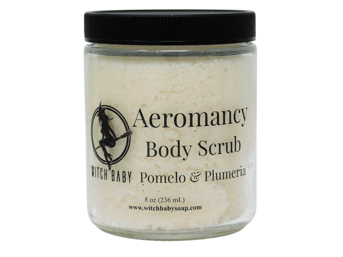 A clear glass jar of Witch Baby Aeromancy Body Scrub with a black lid, containing a light cream-colored scrub, on a white background.