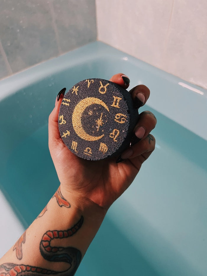 A hand with a snake tattoo holds a round dark blue bath bomb featuring a gold crescent moon and astrological symbols, set against a blue bathtub background.