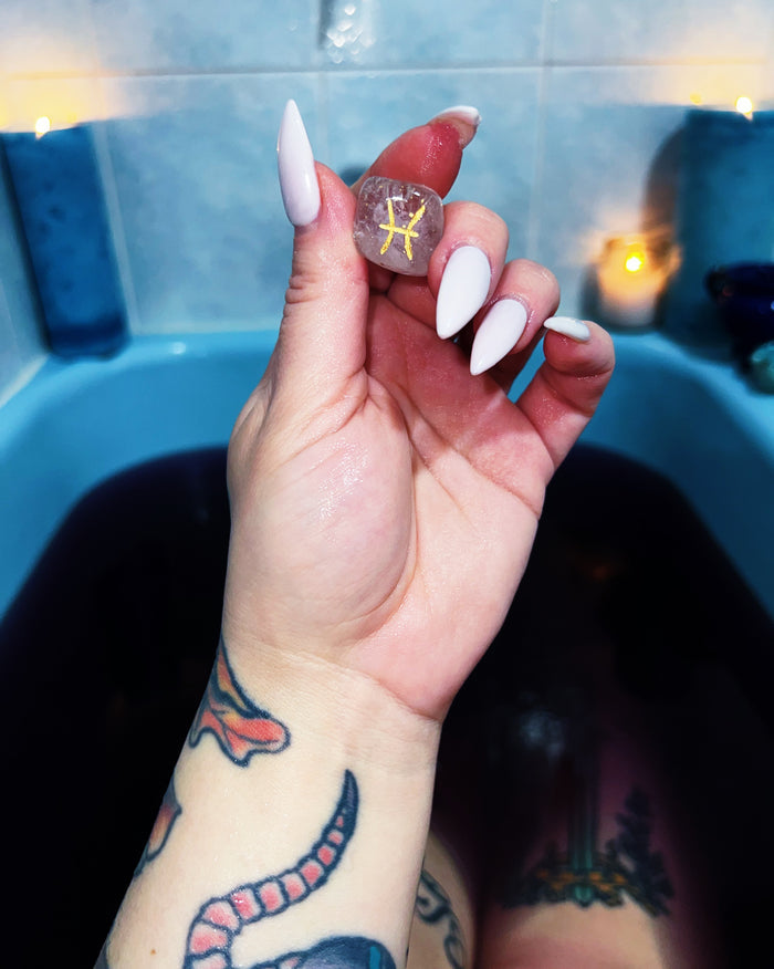 A hand with long white stiletto nails holds a small clear clear quartz featuring a yellow Libra zodiac symbol, set against a blue tiled bathroom background with a bathtub and warm candlelight.
