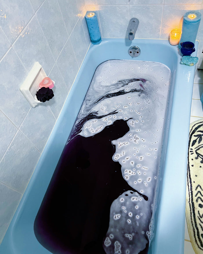 A light blue bathtub filled with dark purple-black liquid and white foam, set in a tiled bathroom with candles and bath accessories around it.