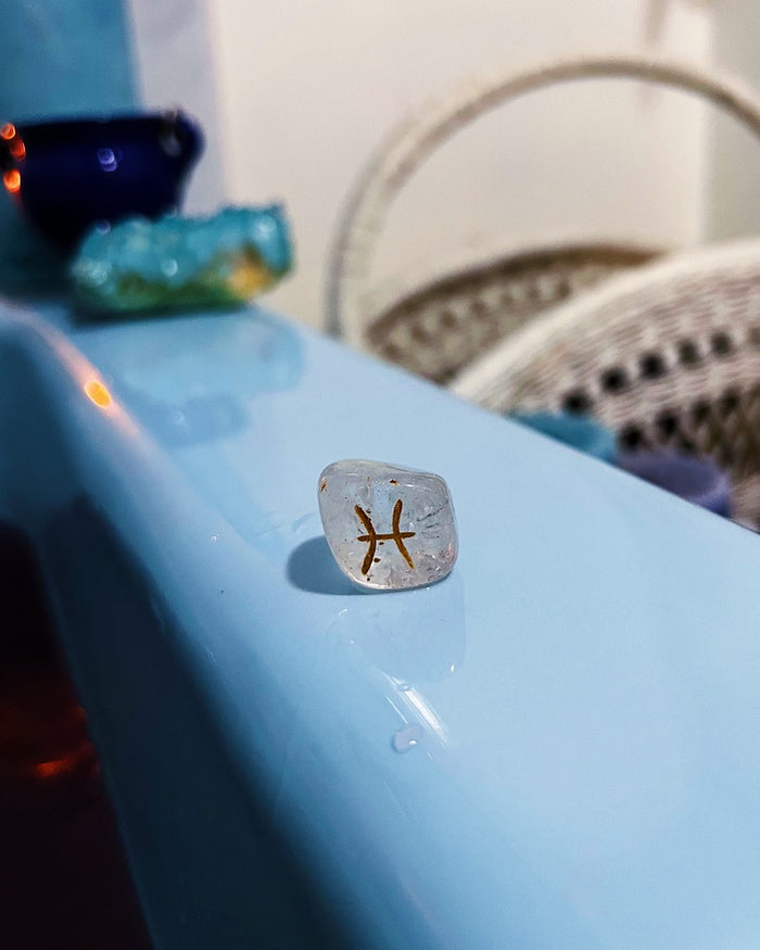 A clear, polished tumbled gemstone with a gold Pisces zodiac symbol inlaid, resting on a light blue surface with a blurred background.