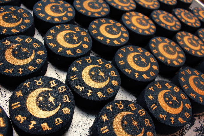 A close-up of multiple round dark blue bath bombs arranged on a light surface, each painted with a gold crescent moon, stars, and zodiac symbols.