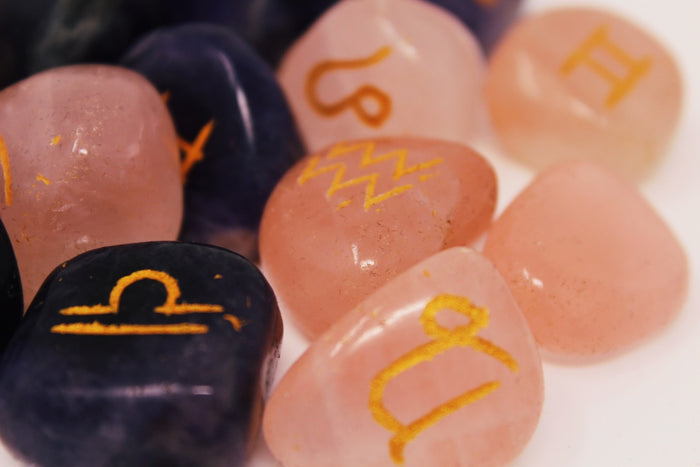 A close-up of polished tumbled gemstones each marked with a small gold zodiac symbol such as Libra, Scorpio, and Aquarius on a white background.