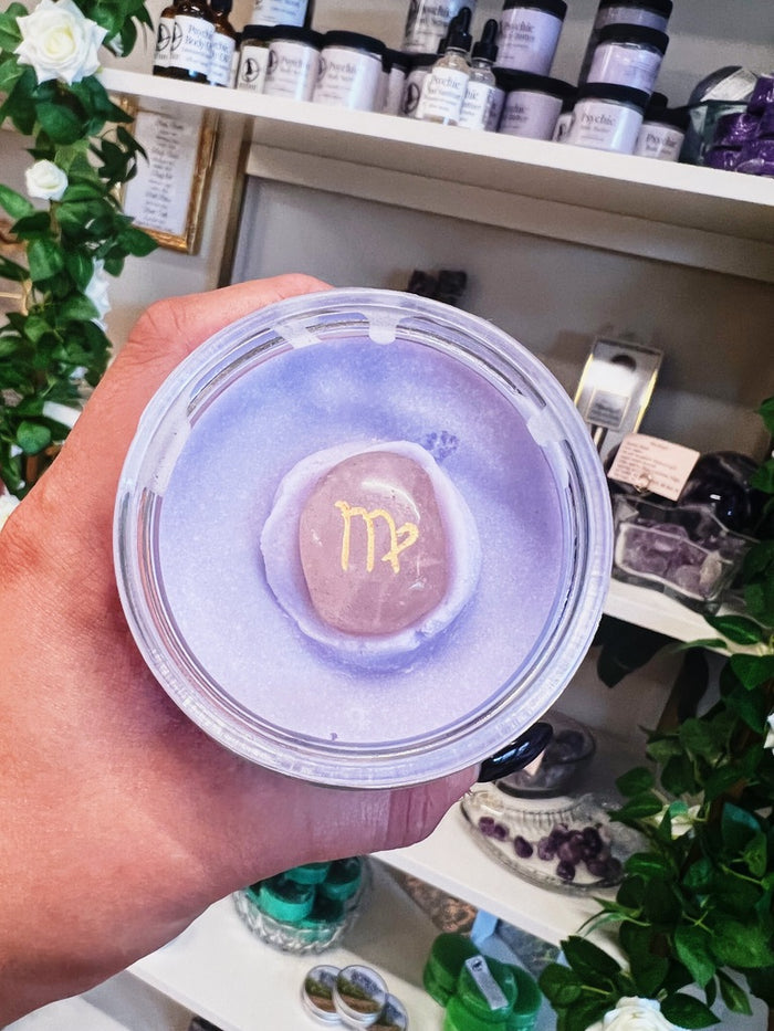 A hand holds a round glass container filled with lavender-colored body butter, featuring an astrology crystal. The background shows shelves filled with various jars and decorative plants.