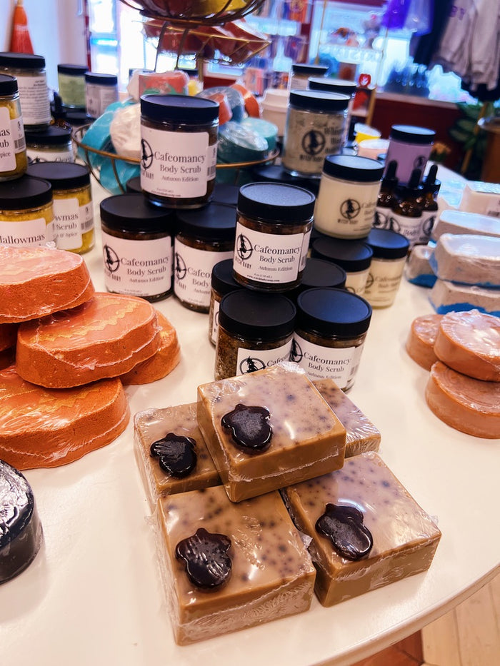 A display of handmade body care products on a white counter, featuring square brown soap bars in the foreground, round orange bath soaps on the left, and multiple glass jars of Cafeomancy body scrubs in the background.
