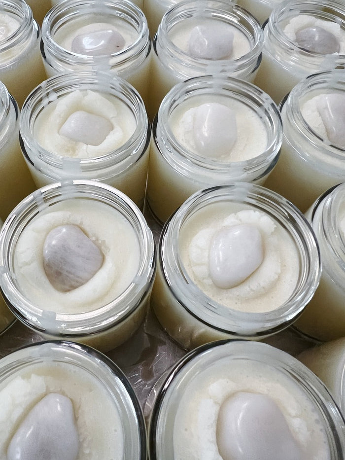 Multiple small clear glass jars filled with a pale creamy white body butter, each topped with a smooth oval white milky quartz crystal, arranged closely together.