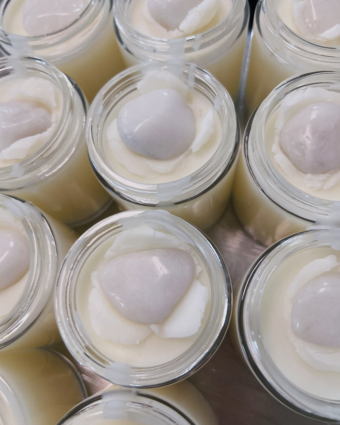 Multiple small clear glass jars filled with a pale creamy white body butter, each topped with a smooth oval white milky quartz crystal, arranged closely together.
