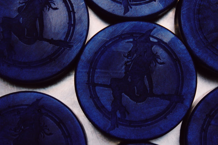 A close-up of several round dark blue soaps featuring an embossed silhouette of a witch riding a broomstick, set against a light background.