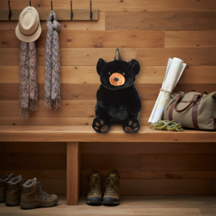 Black bear plush backpack sitting on a wooden mudroom bench surrounded by hiking boots, a canvas duffle bag, and a straw hat