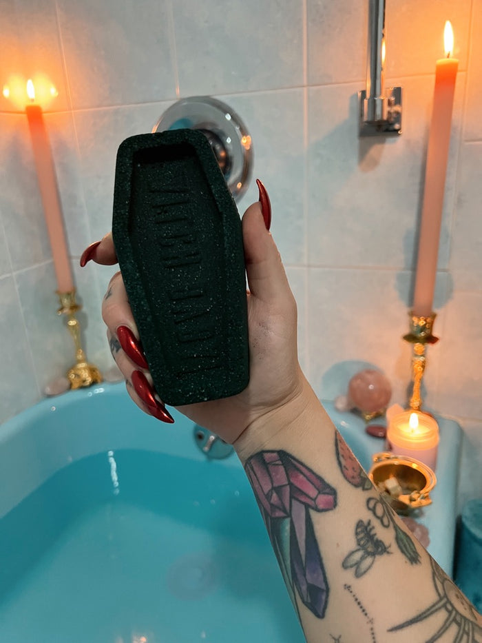 A hand with red nail polish and a tattooed forearm holds a black coffin-shaped bath bomb in front of a lit blue bathtub with candles in the background.