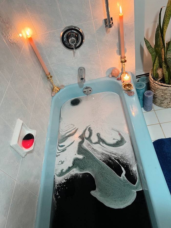 A light blue bathtub filled with black bubbly bath water, surrounded by candles, a soap dish with a red soap, and a potted snake plant in a tiled bathroom.