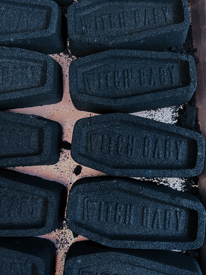 A close-up overhead view of multiple dark gray coffin-shaped bath bombs embossed with the words 'WITCH BABY', arranged on a dusty pink surface with scattered dark powder.