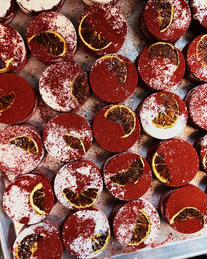 A close-up overhead view of many red round bath bombs arranged on a tray, each topped with a thin dried orange slice and a light dusting of powdered sugar.