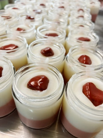 Multiple small clear glass jars filled with layered body butter, each topped with a red jasper crystal, arranged on a metal surface.
