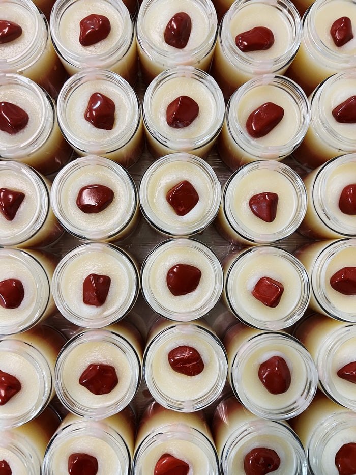 A grid of small clear jars filled with white body butter, each topped with a glossy red jasper gemstone, photographed from above.