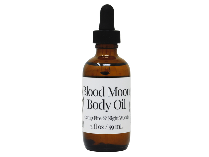 brown glass dropper bottle with white label that reads blood moon  body oil. camp fire & night woods.