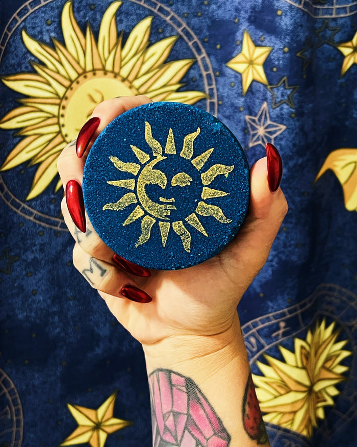 A hand with red nail polish and tattoos holds a round blue glittery bath bomb featuring a gold sun and moon design, set against a dark blue patterned fabric with gold celestial motifs.
