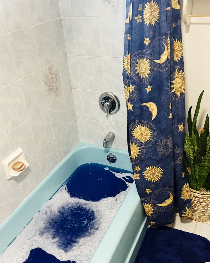 A blue bathtub filled with blue bubbly water in a tiled bathroom, featuring a navy blue shower curtain with a gold celestial pattern of suns, moons, and stars, and a green potted snake plant on the right.