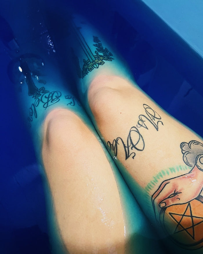 Close-up of two tattooed legs submerged in deep blue water, with black script and illustration tattoos visible on the skin.