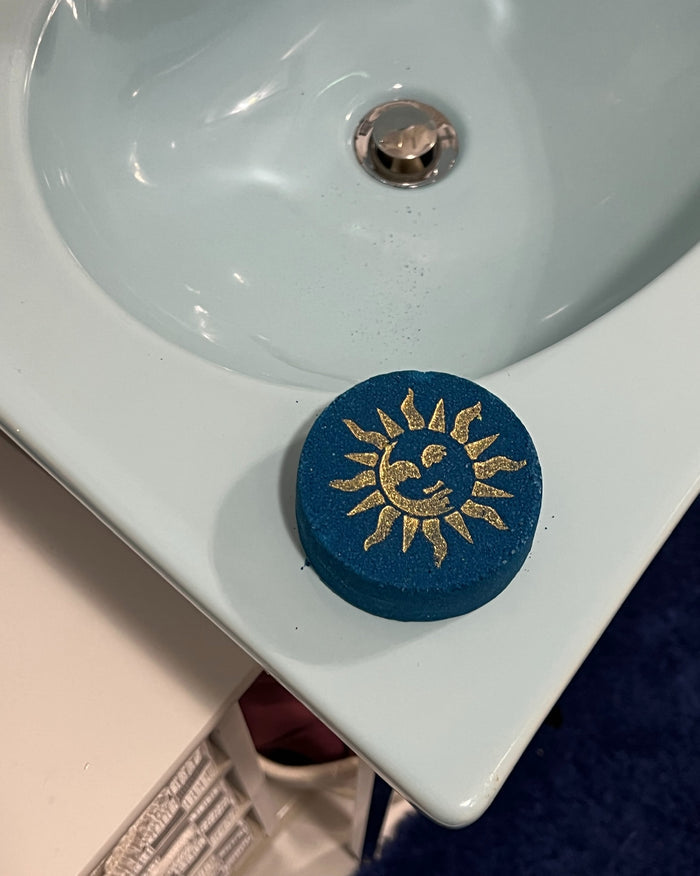 A round blue bath bomb with a gold sun and moon design, resting on the edge of a white ceramic sink.