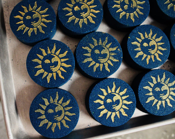 A close-up of several round dark blue bath bombs with a raised gold sun-and-moon airbrush, arranged on a metallic tray.