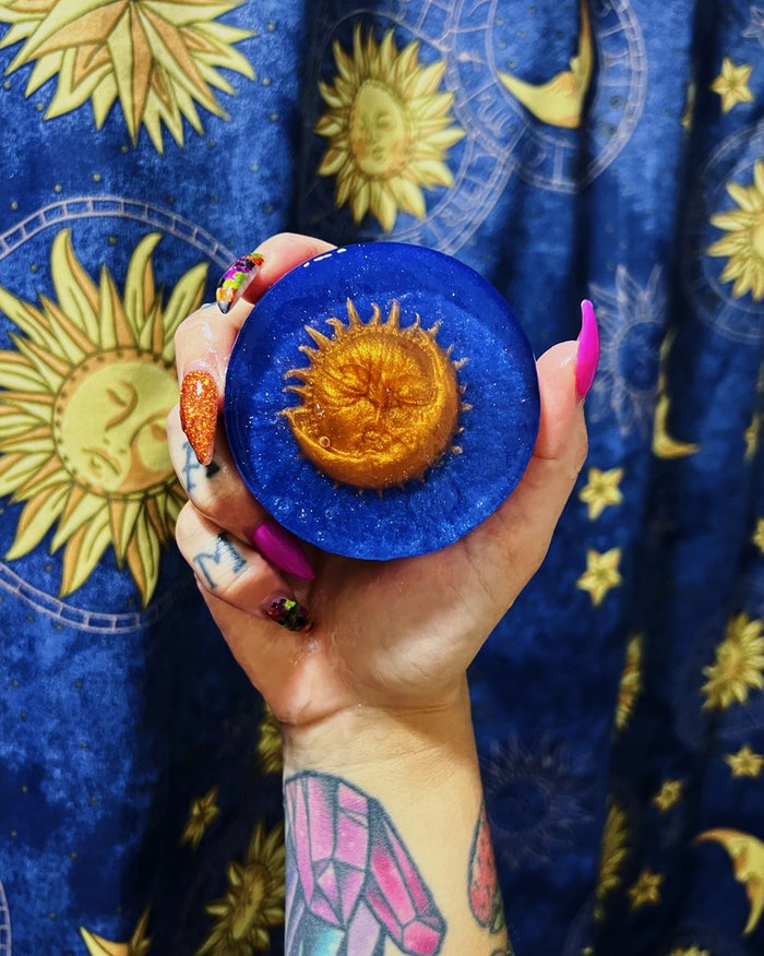 A hand with colorful nail art holds a round blue soap featuring a raised gold sun design in the center, set against a blue fabric backdrop with gold celestial patterns.