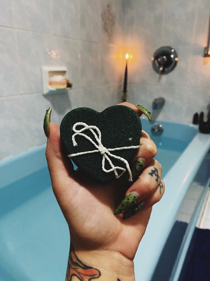 A hand with green glittery nail polish holds a black heart-shaped bath bomb tied with white string, set against a blurred bathroom background with a blue bathtub and a lit candle.