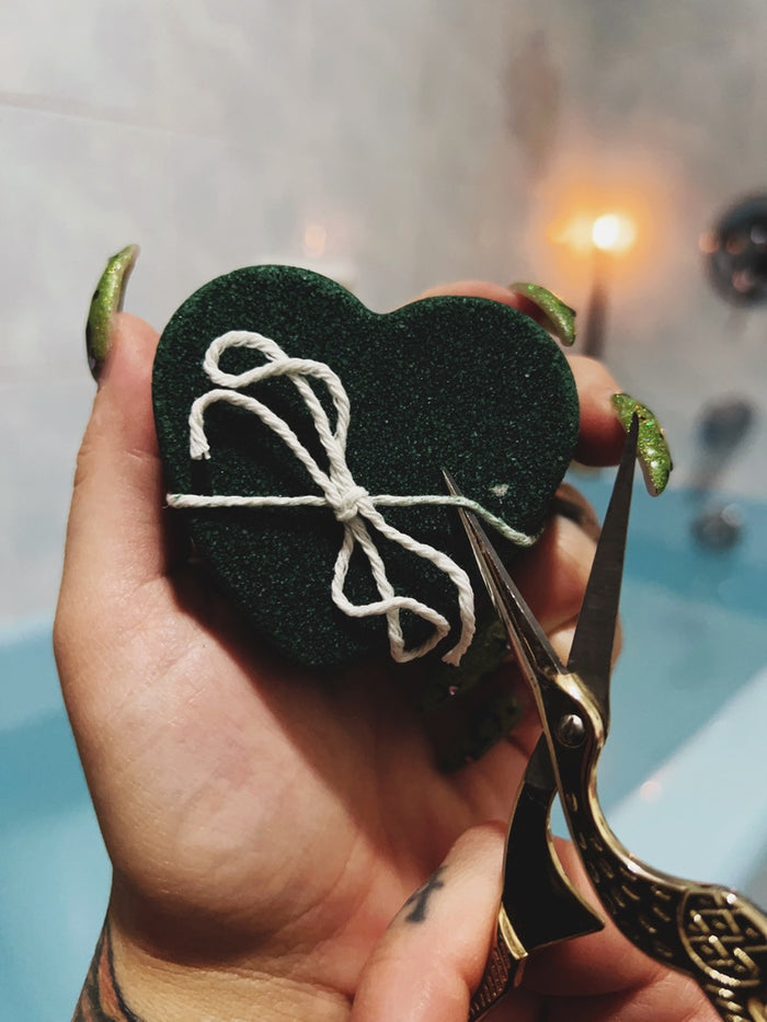 A hand holds a small heart-shaped black bath bombs with white string, while scissors are positioned near it, against a softly blurred bathroom background with a lit candle.