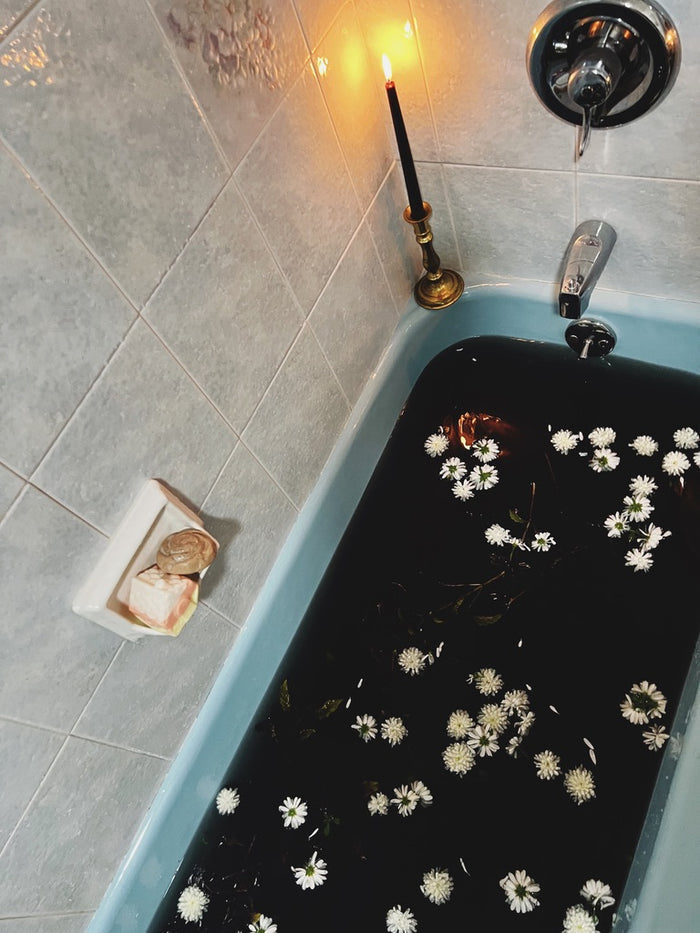 A light blue bathtub filled with black water and floating white daisy-like flowers, set in a tiled bathroom with a lit black candle in a brass holder and a soap dish with soap bars.