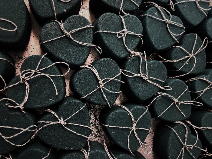 A close-up arrangement of small heart-shaped black bath bombs wrapped in thin beige twine and tied with bows, filling the frame.