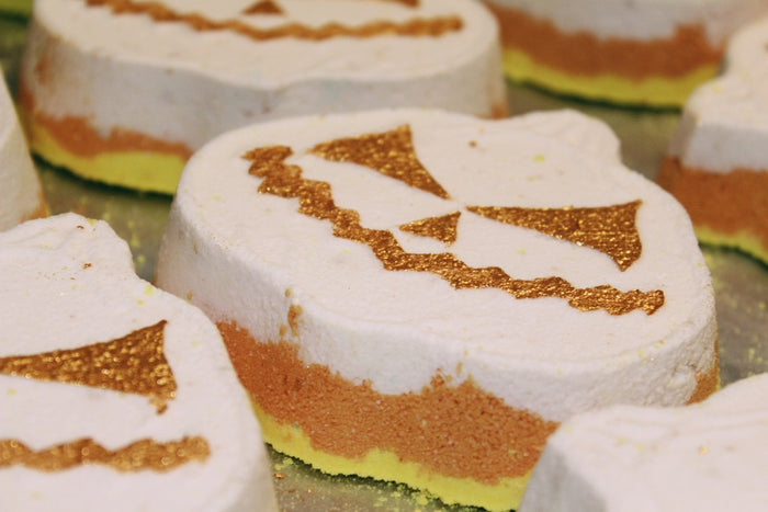 Halloween pumpkin-shaped bath bomb with a white top layer, an orange middle layer, a yellow bottom layer, and a glittery gold jack-o'-lantern face design.