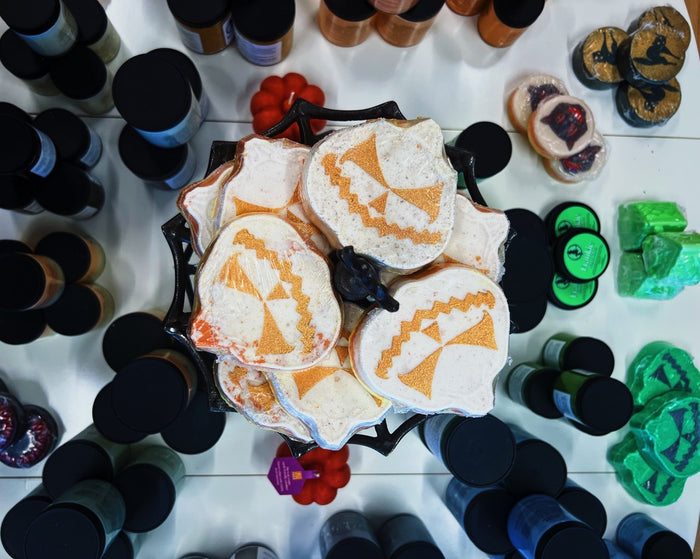A top-down view of a black metal basket filled with white and orange pumpkin-shaped bath bombs, surrounded by many small bottles and colorful bars of soap on a white surface.