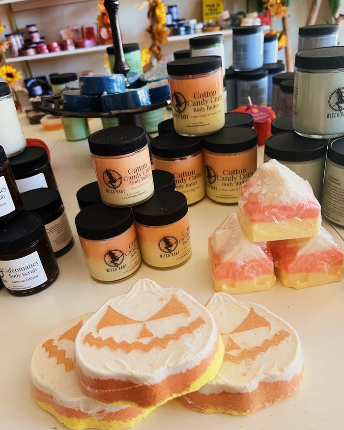 A display of handmade body care products, including pumpkin-shaped soap bars, cotton candy color body lotions in jars, and packaged bath salts on a white counter in a retail setting.