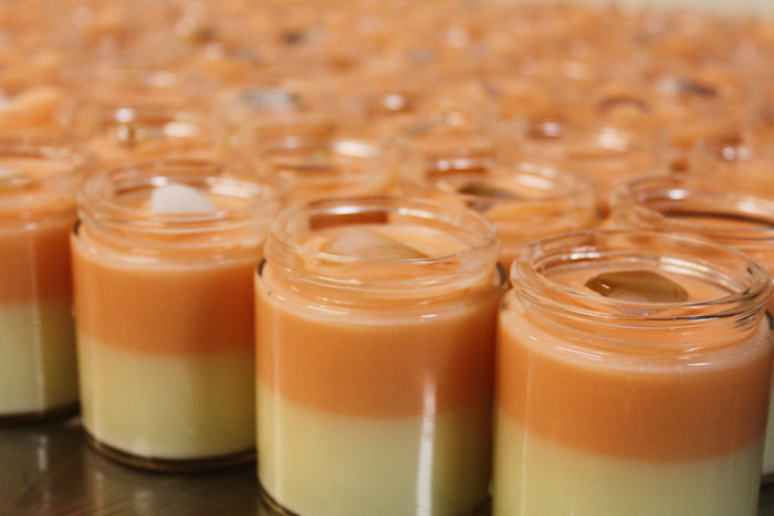 Multiple small clear glass jars filled with a layered body butter, showing a yellow bottom layer and a orange top layer, topped with crystals, arranged closely together.