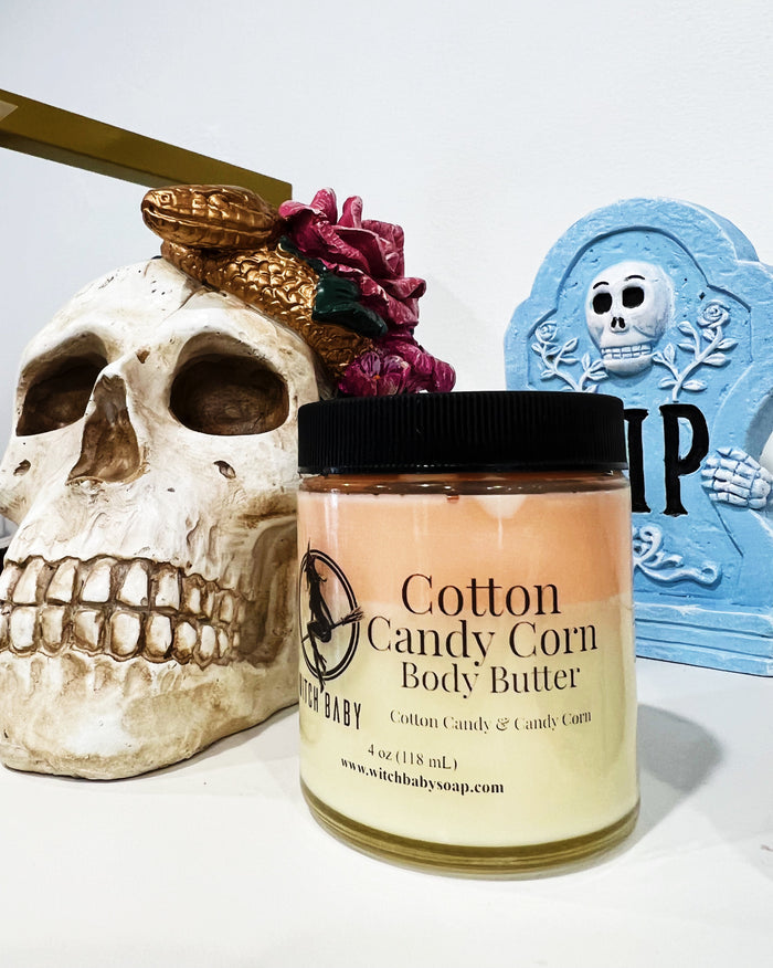 A jar of Cotton Candy Corn Body Butter is displayed on a white surface, styled with a decorative skull, a gold snake, and a blue tombstone prop in the background.