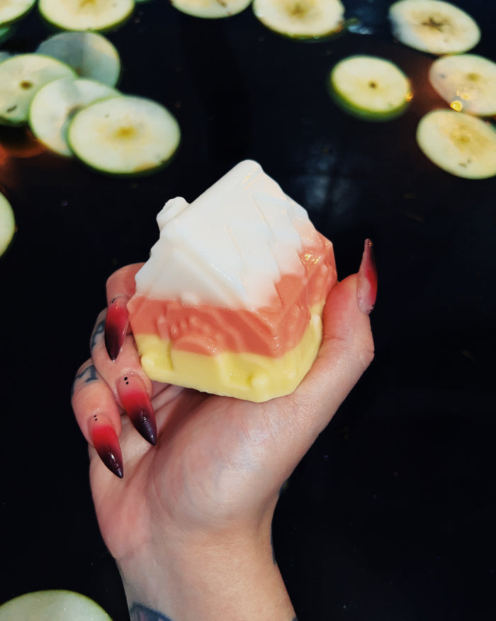 A hand with red-to-black gradient nails holds a triangular layered soap bar featuring white, orange, and yellow candy corn colored sections. The background is a dark surface with floating sliced green apple pieces.