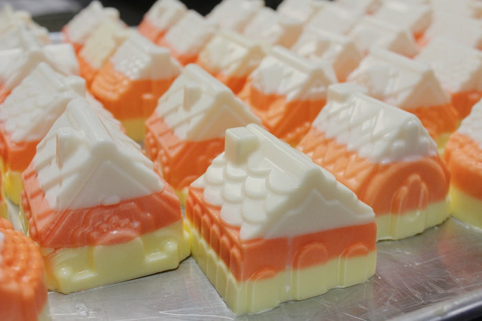 A close-up of several candy corn-shaped soaps arranged on a silver tray. Each chocolate is layered in white, orange, and pale yellow, with the front piece showing a house-like design with a white roof and orange and yellow base.