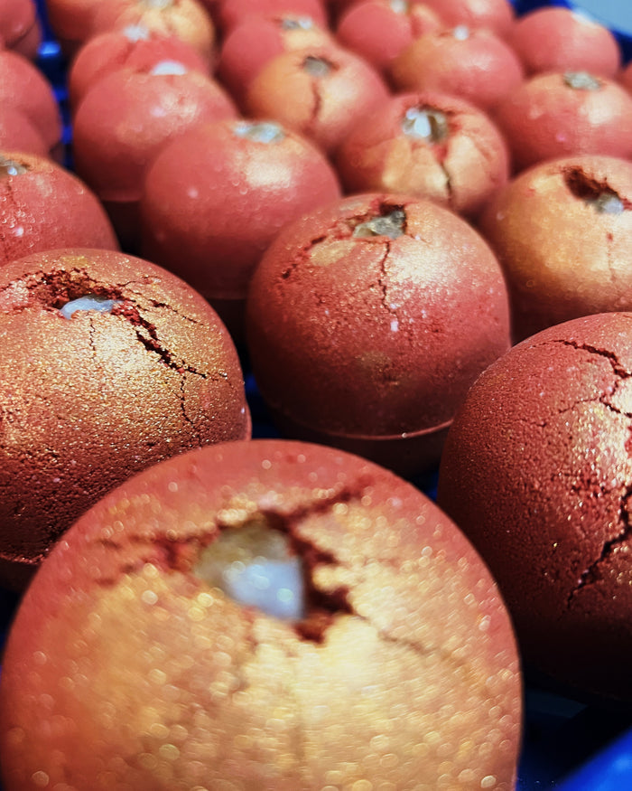 A close-up shot of multiple round, redd bath bombs with a cracked, textured surface, embedded with a citrine crystal, arranged closely together in a tray or container.