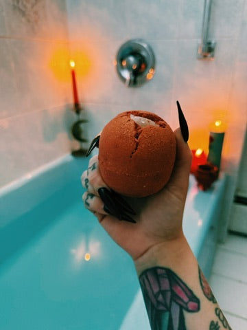 A hand with black fingernails and tattoos holds a red dragons blood bath bomb over a lit blue bathtub with candles in the background.