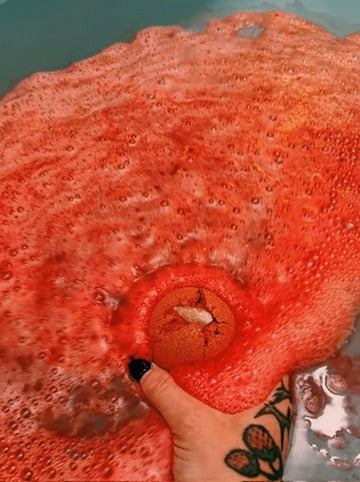 A close-up of a bright orange-red bath bomb or bath salt dissolving in water, creating a large foamy bubble cloud. A tattooed hand with black nail polish is visible at the bottom edge holding the product.