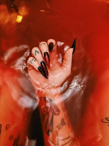 A close-up of a hand with long black stiletto-style nails, partially submerged in red water. The fingers are decorated with black nail wraps and small red accents, and the back of one finger shows the letter 'M' in blue.