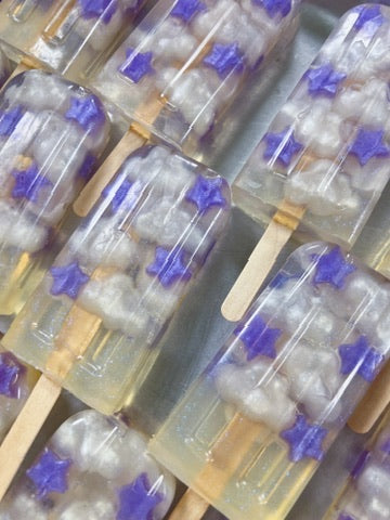 A close-up of several translucent popsicle bar soaps with wooden sticks, filled with white cloud-like pieces and decorated with small purple star accents.