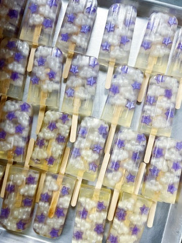 Multiple translucent popsicle-style soaps with wooden sticks are arranged on a metal tray. Each treat contains white pearl-like chunks and small purple star-shaped inclusions.