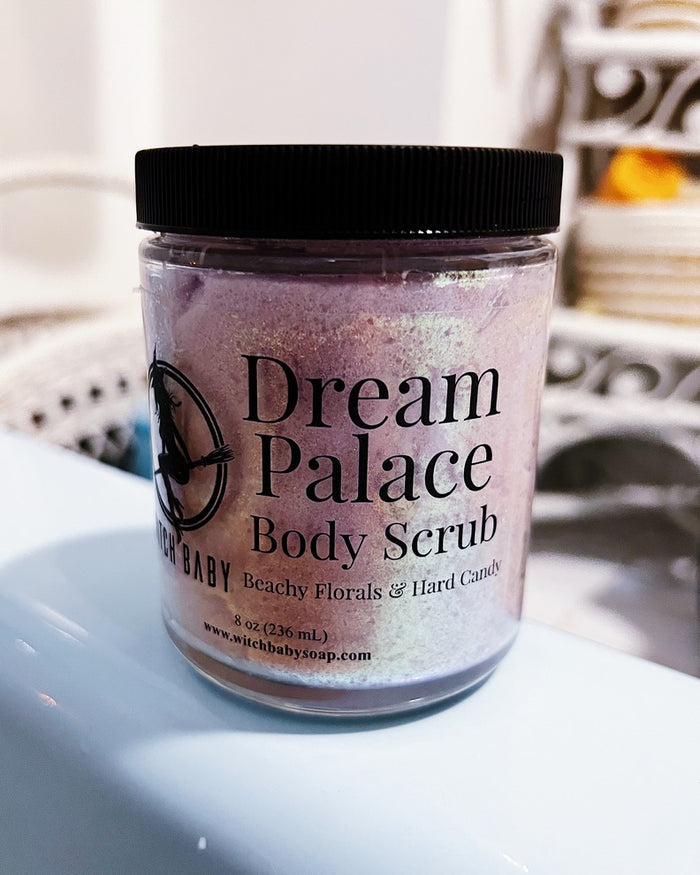 A jar of 'Dream Palace' body scrub by 'Witch Baby Soap' with a black lid and black label, sitting on a white surface with a blurred indoor background.