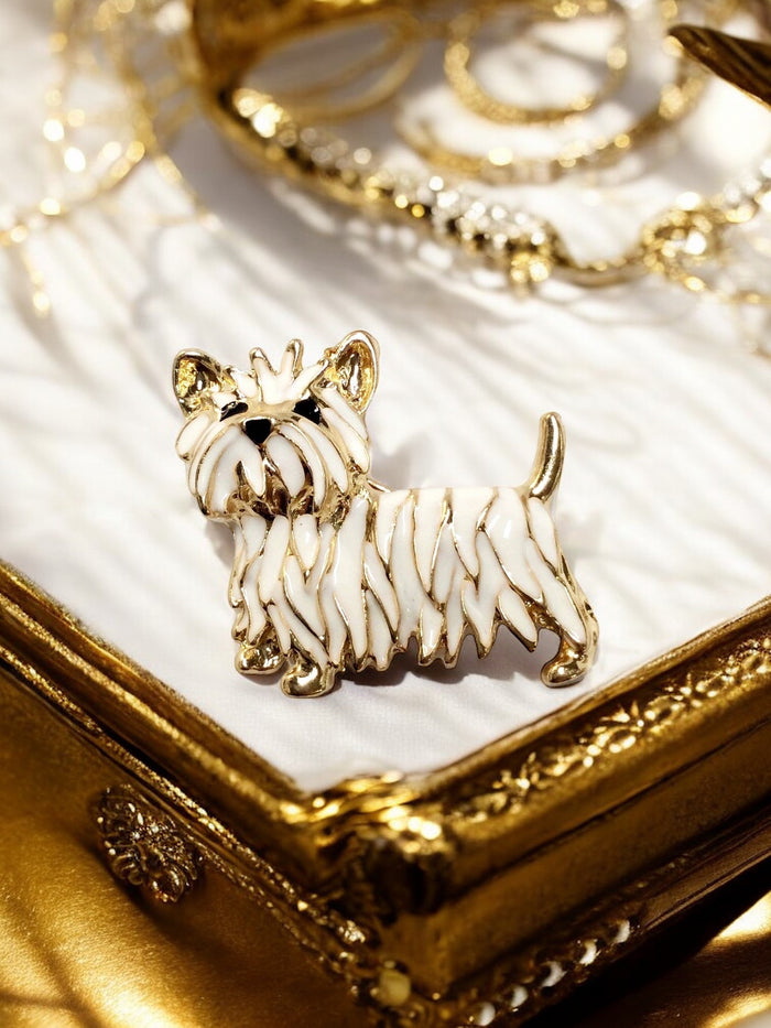 White enamel and gold tone Westie terrier brooch lapel pin displayed on a gold jewelry box with pearl and gold necklaces in the background
