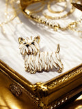White enamel and gold tone Westie terrier brooch lapel pin displayed on a gold jewelry box with pearl and gold necklaces in the background