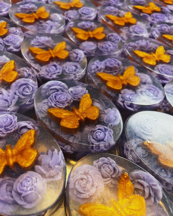 A close-up of multiple oval translucent soaps featuring lavender-purple sculpted roses and orange-gold butterflies inside clear resin.