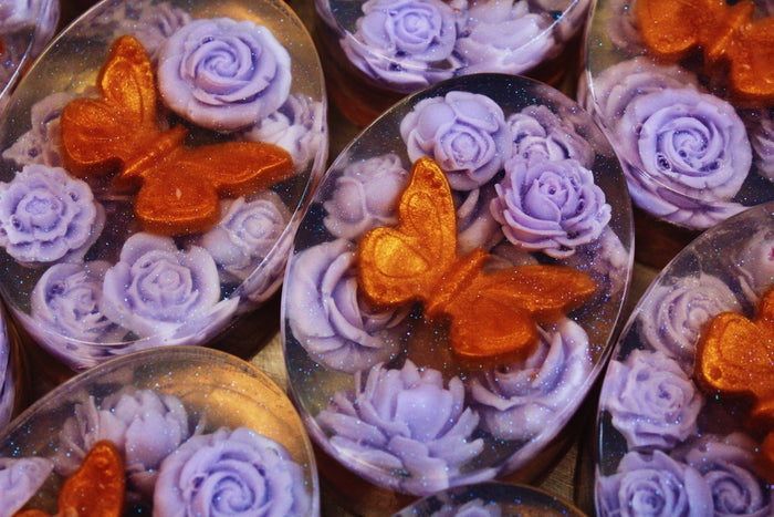 Close-up of oval soaps featuring lavender-purple sculpted roses and glittery orange butterfly motifs.