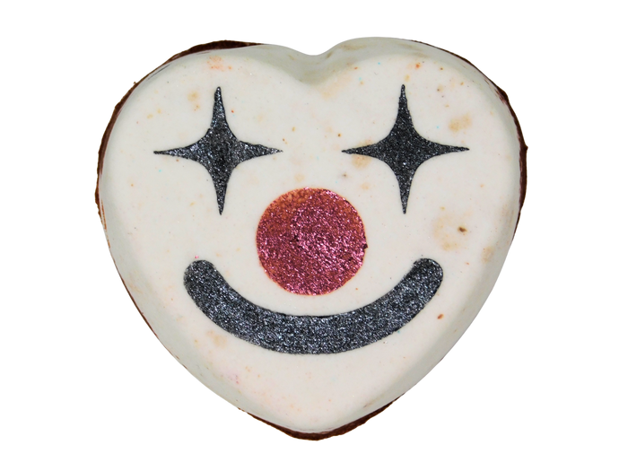 A heart-shaped white bath bomb decorated with a black glittery star-eyed smiley clown face and a glittery red nose, isolated on a white background.