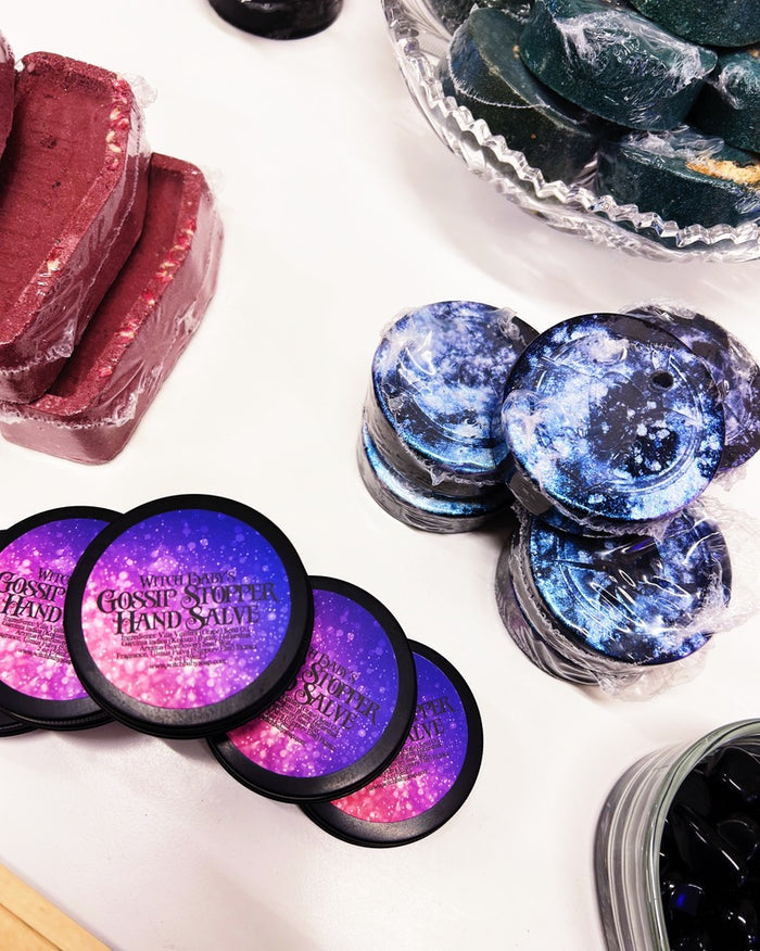 A styled product display of hand salves on a white surface, featuring round tins of 'Gossip Stopper' hand salve with purple and pink galaxy lids, a stack of blue galaxy gossip stopper soaps, and red coffin bath bombs wrapped in clear biodegradable plastic.